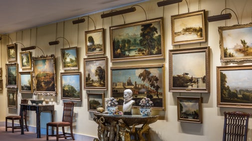 Several paintings on a cream wall, with a small table and wooden chairs in front.